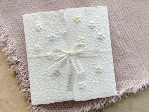 Handgeschöpfte Einladungskarte mit geprägten Blümchen im Gatefold-Design, mit Satinschleife und Büttenrand