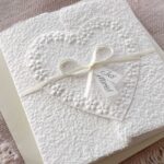 Seitliche Ansicht Glückwunschkarte zur Hochzeit aus handgeschöpftem Papier im Gatefold Design. Motiv ist ein grosses, handgeschöpftes Herz mit Blumenprägung entlang des Randes. Karte und Herz sind weiss in weiss gehalten. Zwei Titelstreifen "Just Married" oder Wunschtitel an einem Schleifchen befestigt, welches die Karte verschliesst.