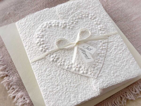 Seitliche Ansicht Glückwunschkarte zur Hochzeit aus handgeschöpftem Papier im Gatefold Design. Motiv ist ein grosses, handgeschöpftes Herz mit Blumenprägung entlang des Randes. Karte und Herz sind weiss in weiss gehalten. Zwei Titelstreifen "Just Married" oder Wunschtitel an einem Schleifchen befestigt, welches die Karte verschliesst.