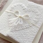 Seitliche Ansicht Hochzeitskarte aus handgeschöpftem Papier im Gatefold Design. Motiv ist ein grosses, handgeschöpftes Herz mit Blumenprägung entlang des Randes. Karte und Herz sind weiss in weiss gehalten. Zwei Titelstreifen "Just Married" oder Wunschtitel an einem Schleifchen befestigt, welches die Karte verschliesst.