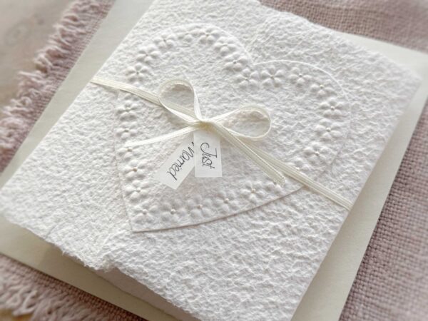 Seitliche Ansicht Hochzeitskarte aus handgeschöpftem Papier im Gatefold Design. Motiv ist ein grosses, handgeschöpftes Herz mit Blumenprägung entlang des Randes. Karte und Herz sind weiss in weiss gehalten. Zwei Titelstreifen "Just Married" oder Wunschtitel an einem Schleifchen befestigt, welches die Karte verschliesst.