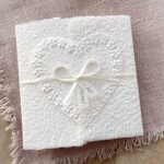 Hochzeitskarte aus handgeschöpftem Papier im Gatefold Design. Motiv ist ein grosses, handgeschöpftes Herz mit Blumenprägung entlang des Randes. Karte und Herz sind weiss in weiss gehalten. Zwei Titelstreifen "Just Married" oder Wunschtitel an einem Schleifchen befestigt, welches die Karte verschliesst.