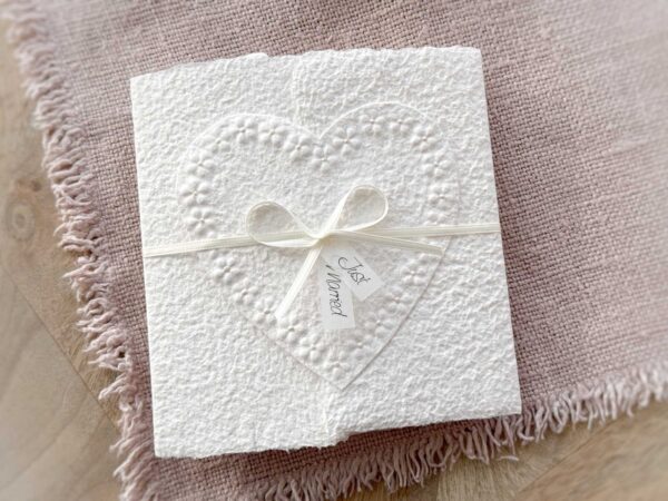 Hochzeitskarte aus handgeschöpftem Papier im Gatefold Design. Motiv ist ein grosses, handgeschöpftes Herz mit Blumenprägung entlang des Randes. Karte und Herz sind weiss in weiss gehalten. Zwei Titelstreifen "Just Married" oder Wunschtitel an einem Schleifchen befestigt, welches die Karte verschliesst.