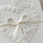 Nahaufnahme Glückwunschkarte zur Hochzeit aus handgeschöpftem Papier im Gatefold Design. Motiv ist ein grosses, handgeschöpftes Herz mit Blumenprägung entlang des Randes. Karte und Herz sind weiss in weiss gehalten. Zwei Titelstreifen "Just Married" oder Wunschtitel an einem Schleifchen befestigt, welches die Karte verschliesst.