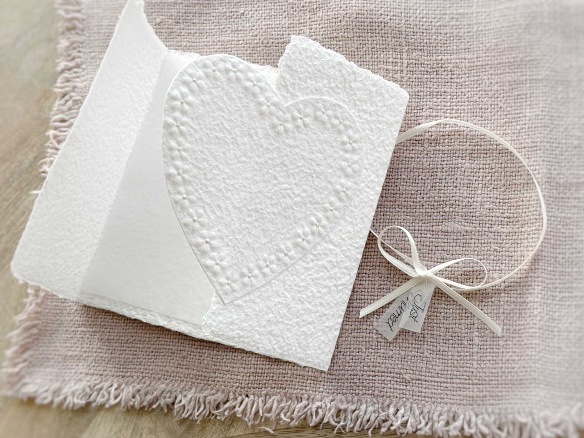 Geöffnete der Glückwunschkarte zur Hochzeit aus handgeschöpftem Papier im Gatefold Design. Motiv ist ein grosses, handgeschöpftes Herz mit Blumenprägung entlang des Randes. Karte und Herz sind weiss in weiss gehalten. Zwei Titelstreifen "Just Married" oder Wunschtitel an einem Schleifchen befestigt, welches die Karte verschliesst.