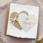 Glückwunschkarte zur Hochzeit aus handgeschöpftem Papier im Gatefold Design. Mit einem Doppelherz aus zwei verschiedenen beige-tönen und Titel "Just Married". Verschluss mit einem Satinband in creme.