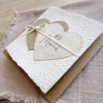 Seitliche Ansicht Glückwunschkarte zur Hochzeit aus handgeschöpftem Papier im Gatefold Design. Mit einem Doppelherz aus zwei verschiedenen beige-tönen und Titel "Just Married". Verschluss mit einem Satinband in creme.