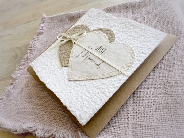 Seitliche Ansicht Glückwunschkarte zur Hochzeit aus handgeschöpftem Papier im Gatefold Design. Mit einem Doppelherz aus zwei verschiedenen beige-tönen und Titel "Just Married". Verschluss mit einem Satinband in creme.