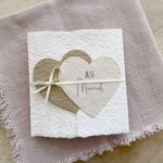 Glückwunschkarte zur Hochzeit aus handgeschöpftem Papier im Gatefold Design. Mit einem Doppelherz aus zwei verschiedenen beige-tönen und Titel "Just Married". Verschluss mit einem Satinband in creme.