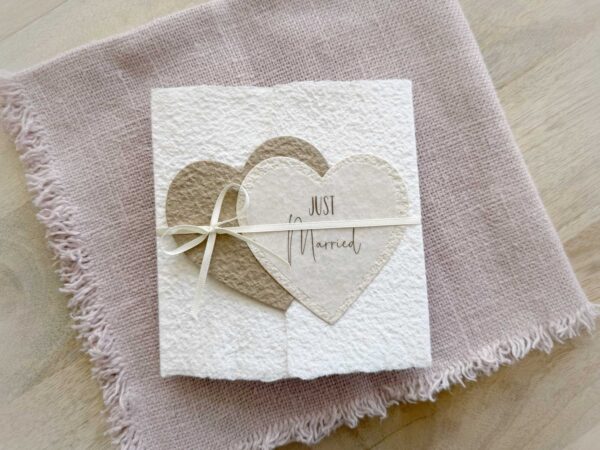 Glückwunschkarte zur Hochzeit aus handgeschöpftem Papier im Gatefold Design. Mit einem Doppelherz aus zwei verschiedenen beige-tönen und Titel "Just Married". Verschluss mit einem Satinband in creme.