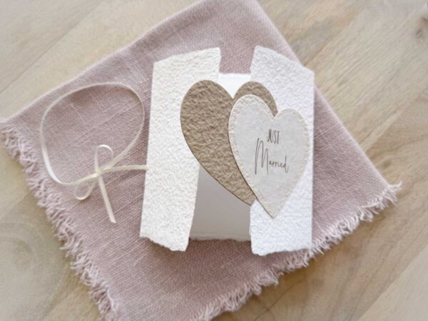 Geöffnete Ansicht Glückwunschkarte zur Hochzeit aus handgeschöpftem Papier im Gatefold Design. Mit einem Doppelherz aus zwei verschiedenen beige-tönen und Titel "Just Married". Verschluss mit einem Satinband in creme.
