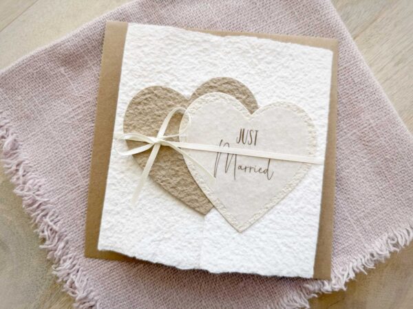 Glückwunschkarte zur Hochzeit aus handgeschöpftem Papier im Gatefold Design. Mit einem Doppelherz aus zwei verschiedenen beige-tönen und Titel "Just Married". Verschluss mit einem Satinband in creme.