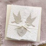 Glückwunschkarte zur Hochzeit aus handgeschöpftem Papier im Gatefold Design. Weisse Karte mit beigen Tauben, die aufeinander zu fliegen. Kleines Herz mit dem Titel "Just Married". Verschluss mit einem Satinband in creme.