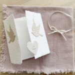 Geöffnete Glückwunschkarte zur Hochzeit aus handgeschöpftem Papier im Gatefold Design. Weisse Karte mit beigen Tauben, die aufeinander zu fliegen. Kleines Herz mit dem Titel "Just Married". Verschluss mit einem Satinband in creme.