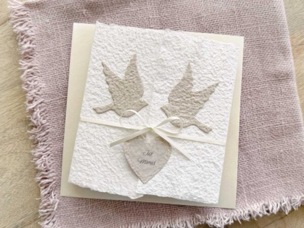 Glückwunschkarte zur Hochzeit aus handgeschöpftem Papier im Gatefold Design. Weisse Karte mit beigen Tauben, die aufeinander zu fliegen. Kleines Herz mit dem Titel "Just Married". Verschluss mit einem Satinband in creme.