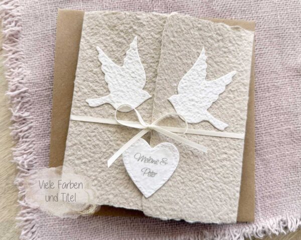 Personalisierte Glückwunschkarte zur Hochzeit aus handgeschöpftem Papier im Gatefold Design. Beige Karte mit weissen Tauben, die aufeinander zu fliegen. Kleines Herz mit Namen des Brautpaars. Verschluss mit einem Satinband in creme.