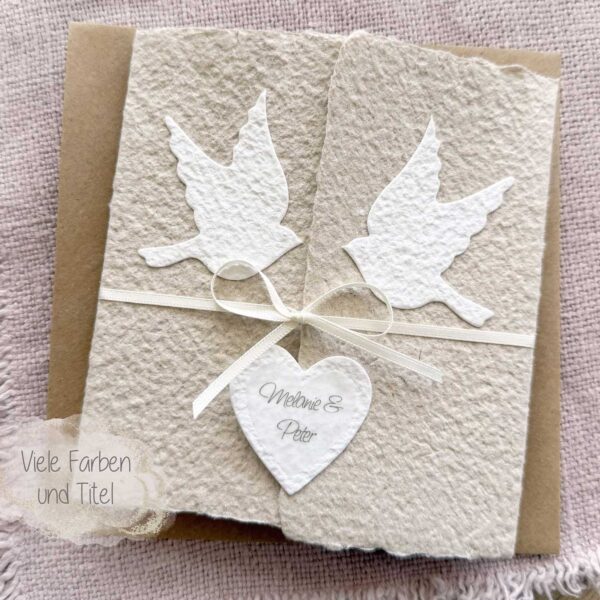 Personalisierte Glückwunschkarte zur Hochzeit aus handgeschöpftem Papier im Gatefold Design. Beige Karte mit weissen Tauben, die aufeinander zu fliegen. Kleines Herz mit Namen des Brautpaars. Verschluss mit einem Satinband in creme.