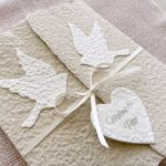 Nahaufnahme personalisierte Glückwunschkarte zur Hochzeit aus handgeschöpftem Papier im Gatefold Design. Beige Karte mit weissen Tauben, die aufeinander zu fliegen. Kleines Herz mit Namen des Brautpaars. Verschluss mit einem Satinband in creme.