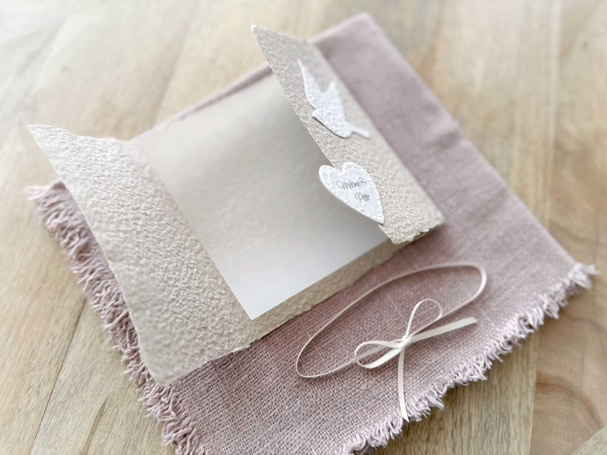 Geöffnete Ansicht der personalisierten Glückwunschkarte zur Hochzeit aus handgeschöpftem Papier im Gatefold Design. Beige Karte mit weissen Tauben, die aufeinander zu fliegen. Kleines Herz mit Namen des Brautpaars. Verschluss mit einem Satinband in creme.