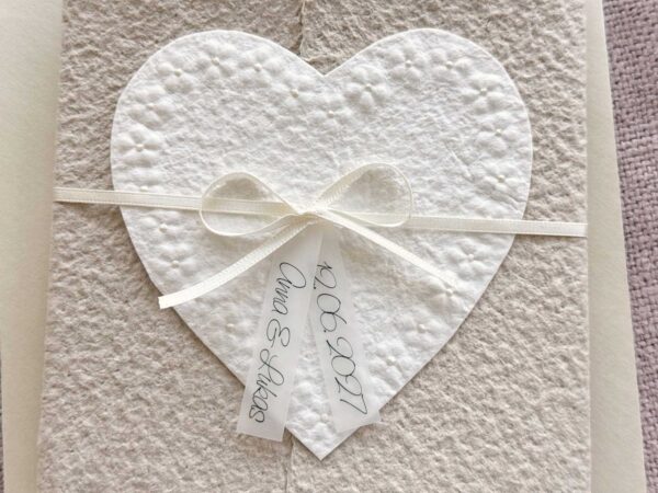 Nahaufnahme personalisierte Hochzeitseinladung Blumenherz aus handgeschöpftem Büttenpapier mit floraler Herzprägung und Titelstreifen am SatinbandPersonalisierte Hochzeitseinladung Blumenherz aus handgeschöpftem Büttenpapier mit floraler Herzprägung und Titelstreifen am Satinband