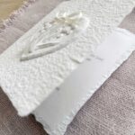 Leicht aufgeklappte Hochzeitseinladung "Büttenherz" aus handgeschöpftem Papier weiss in weiss. Mit personalisiertem Titel auf zwei transparentpapierstreifen.