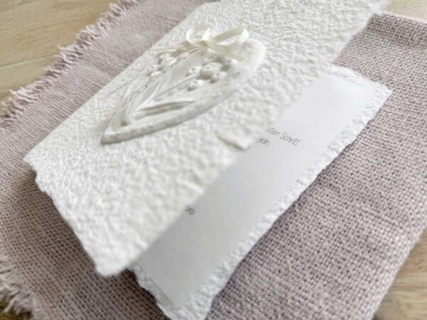 Leicht aufgeklappte Hochzeitseinladung "Büttenherz" aus handgeschöpftem Papier weiss in weiss. Mit personalisiertem Titel auf zwei transparentpapierstreifen.