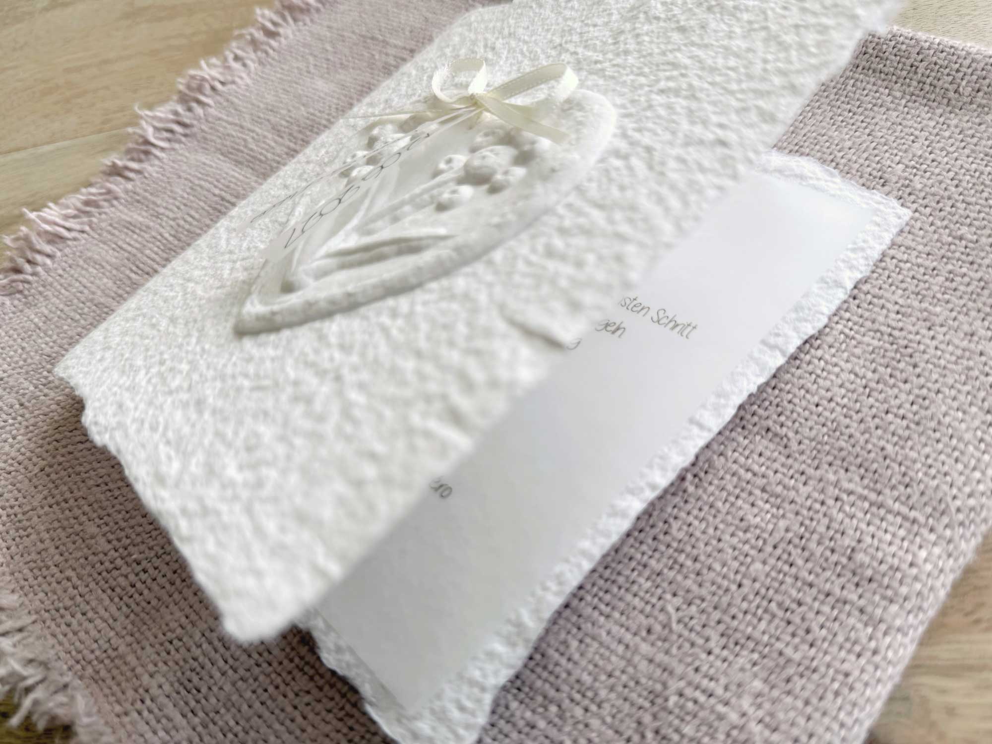 Leicht aufgeklappte Hochzeitseinladung "Büttenherz" aus handgeschöpftem Papier weiss in weiss. Mit personalisiertem Titel auf zwei transparentpapierstreifen.