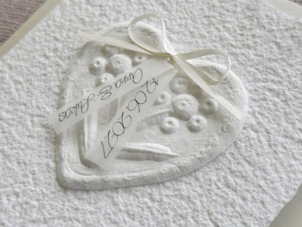 Nahaufnahme Hochzeitseinladung "Büttenherz" aus handgeschöpftem Papier weiss in weiss. Mit personalisiertem Titel auf zwei transparentpapierstreifen.
