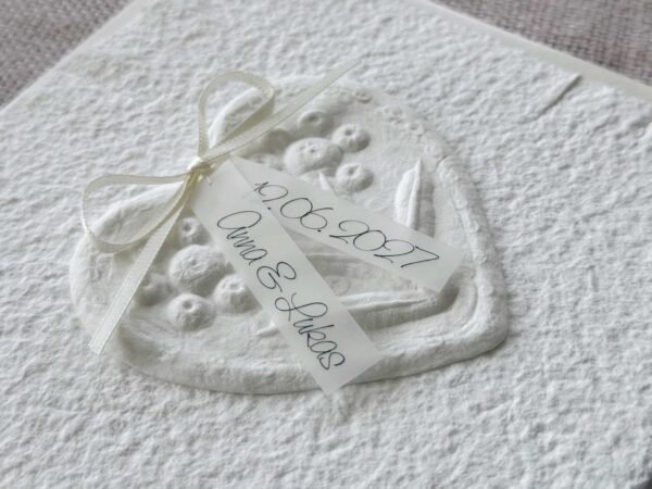 Seitliche Nahaufnahme Hochzeitseinladung "Büttenherz" aus handgeschöpftem Papier weiss in weiss. Mit personalisiertem Titel auf zwei transparentpapierstreifen.