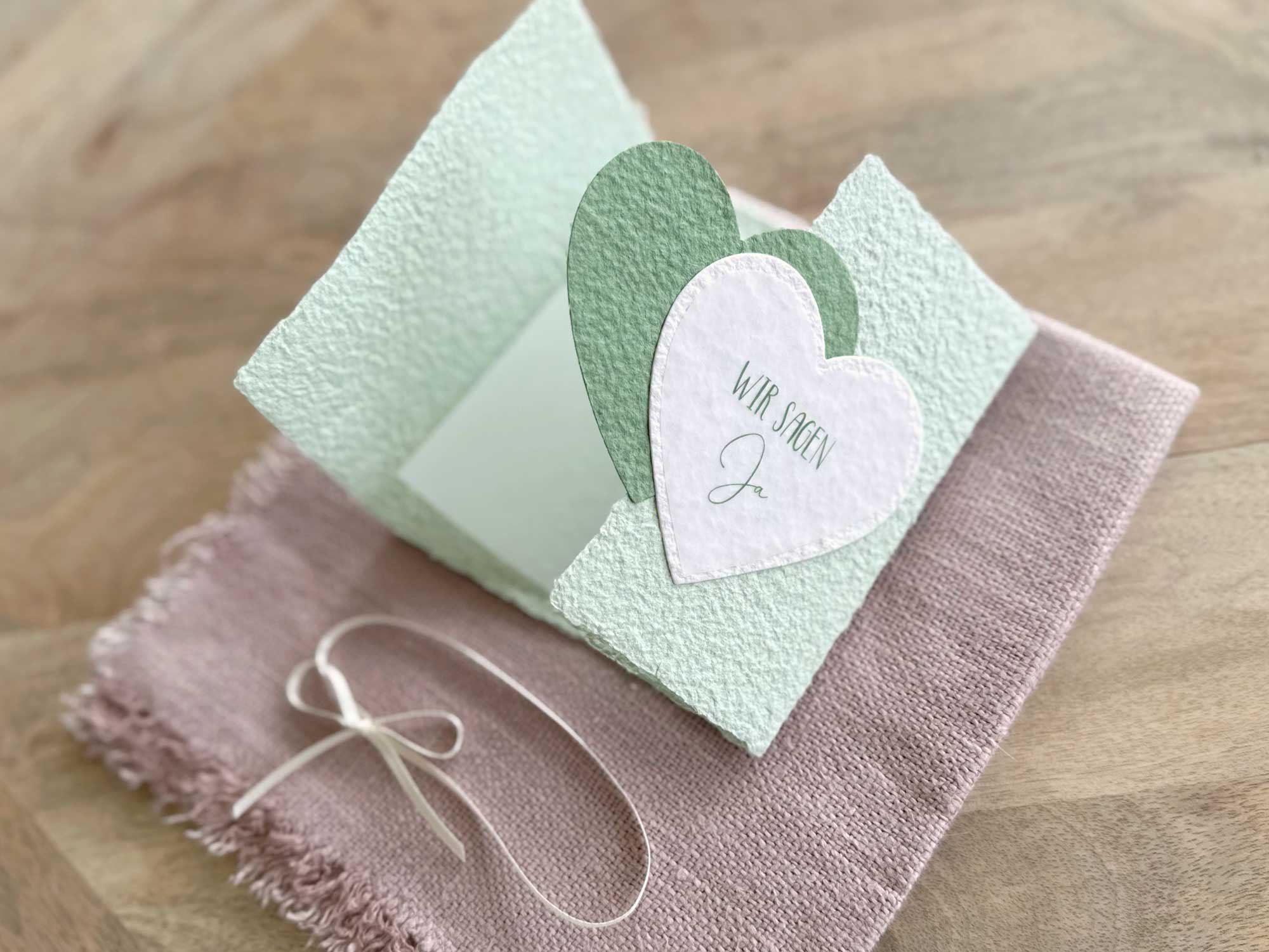 Aufgeklappte Gatefold-Hochzeitseinladung aus handgeschöpftem Papier in zartem Grün mit zwei herzen in dunkelgrün und weiss. Titel "Wir sagen Ja".
