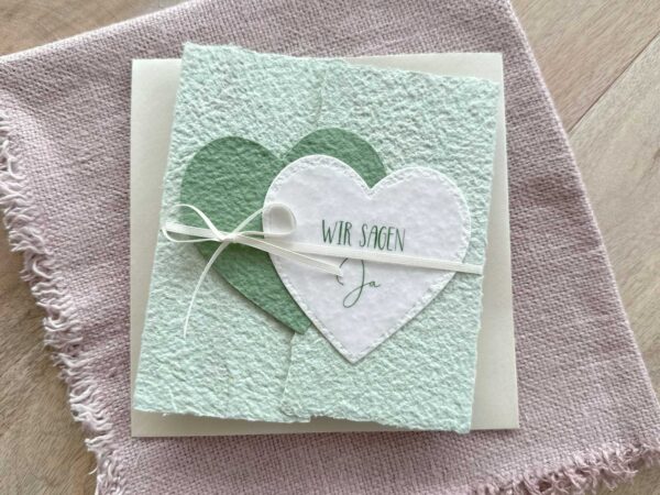 Personalisierbare Gatefold-Hochzeitseinladung aus handgeschöpftem Papier in zartem Grün mit zwei herzen in dunkelgrün und weiss. Titel "Wir sagen Ja".