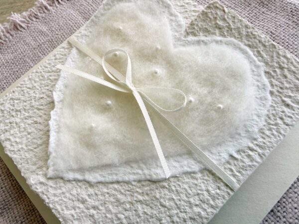 Nahaufnahme Hochzeitseinladung aus handgeschöpftem Papier mit edlem, handgefilztem Perlenherz als Motiv. Gatefold Design mit Satinband verschlossen.