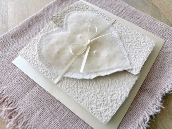 Seitliche Ansicht Hochzeitseinladung aus handgeschöpftem Papier mit edlem, handgefilztem Perlenherz als Motiv. Gatefold Design mit Satinband verschlossen.