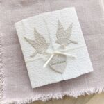 Besondere Hochzeitseinladung «Tauben» aus handgeschöpftem Büttenpapier im Altarfalz Desing mit zwei beigen Tauben und elegantem Satinband. Personalisierter Titel auf beigem Herz.