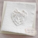 Personalisierte Glückwunschkarte zur Hochzeit aus handgeschöpftem Papier. Mit einem 3D-Herz aus einem Springerle Herz, welches zwei Blumen aus einer Wurzel zeigt. In der edlen Hochzeitskombination "weiss in weiss".
