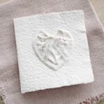 Glückwunschkarte zur Hochzeit aus handgeschöpftem Papier. Mit einem 3D-Herz aus einem Springerle Herz, welches zwei Blumen aus einer Wurzel zeigt. In der edlen Hochzeitskombination "weiss in weiss".