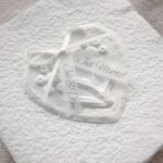 Nahaufnahme personalisierte Glückwunschkarte zur Hochzeit aus handgeschöpftem Papier. Mit einem 3D-Herz aus einem Springerle Herz, welches zwei Blumen aus einer Wurzel zeigt. In der edlen Hochzeitskombination "weiss in weiss".