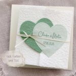 Personalisierte Glückwunschkarte zur Hochzeit aus handgeschöpftem Papier im Gatefold Design. Mit einem Doppelherz aus zwei verschiedenen grün-tönen und einem Wunschtitel versehen. Verschluss mit einem Satinband in creme mit Goldkante.