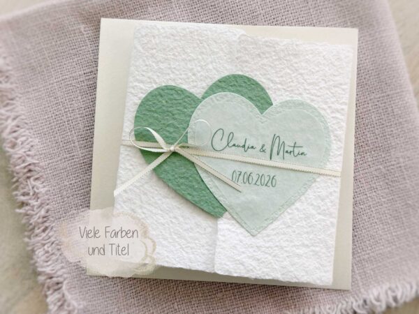 Personalisierte Glückwunschkarte zur Hochzeit aus handgeschöpftem Papier im Gatefold Design. Mit einem Doppelherz aus zwei verschiedenen grün-tönen und einem Wunschtitel versehen. Verschluss mit einem Satinband in creme mit Goldkante.
