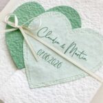 Nahaufnahme personalisierte Glückwunschkarte zur Hochzeit aus handgeschöpftem Papier im Gatefold Design. Mit einem Doppelherz aus zwei verschiedenen grün-tönen und einem Wunschtitel versehen. Verschluss mit einem Satinband in creme mit Goldkante.