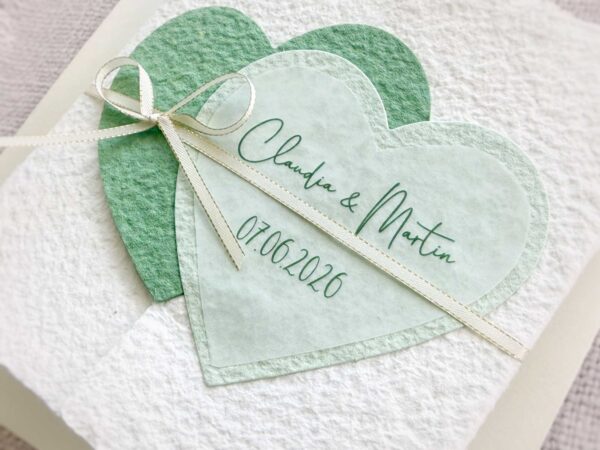 Nahaufnahme personalisierte Glückwunschkarte zur Hochzeit aus handgeschöpftem Papier im Gatefold Design. Mit einem Doppelherz aus zwei verschiedenen grün-tönen und einem Wunschtitel versehen. Verschluss mit einem Satinband in creme mit Goldkante.