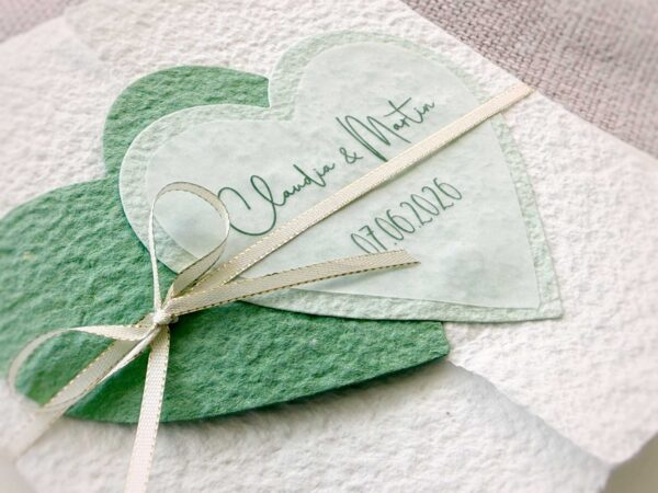 Seitliche Nahaufnahme der personalisierten Glückwunschkarte zur Hochzeit aus handgeschöpftem Papier im Gatefold Design. Mit einem Doppelherz aus zwei verschiedenen grün-tönen und einem Wunschtitel versehen. Verschluss mit einem Satinband in creme mit Goldkante.