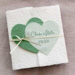 Gatefold Glückwunschkarte zur Hochzeit aus handgeschöpftem Papier. Mit einem Doppelherz aus zwei verschiedenen grün-tönen und einem Wunschtitel versehen. Verschluss mit einem Satinband in creme mit Goldkante.