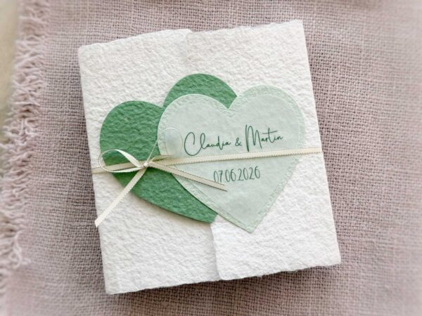 Gatefold Glückwunschkarte zur Hochzeit aus handgeschöpftem Papier. Mit einem Doppelherz aus zwei verschiedenen grün-tönen und einem Wunschtitel versehen. Verschluss mit einem Satinband in creme mit Goldkante.