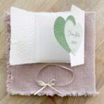 Geöffnete Gatefold Glückwunschkarte zur Hochzeit aus handgeschöpftem Papier. Mit einem Doppelherz aus zwei verschiedenen grün-tönen und einem Wunschtitel versehen. Verschluss mit einem Satinband in creme mit Goldkante.