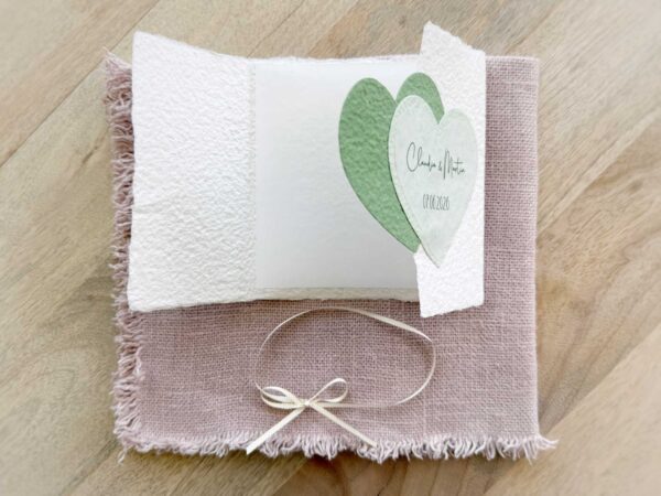 Geöffnete Gatefold Glückwunschkarte zur Hochzeit aus handgeschöpftem Papier. Mit einem Doppelherz aus zwei verschiedenen grün-tönen und einem Wunschtitel versehen. Verschluss mit einem Satinband in creme mit Goldkante.