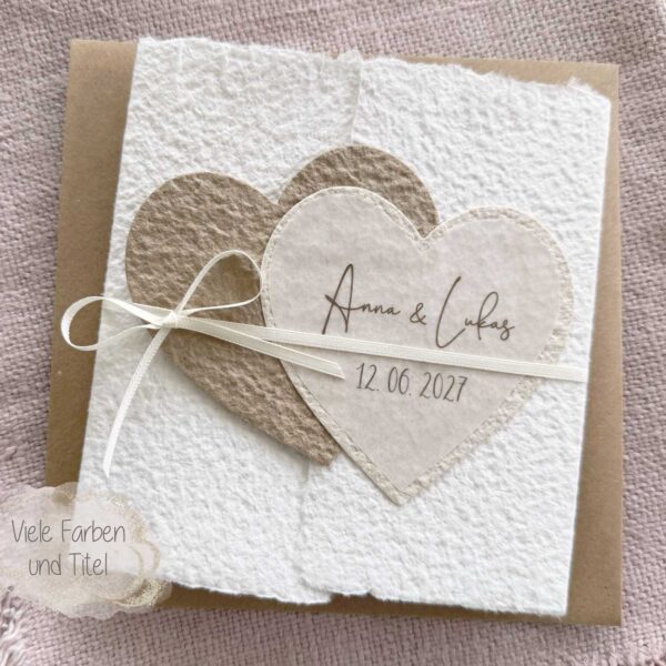 Personalisierte Hochzeitseinladung aus handgeschöpftem Papier im Gatefold Design. Motiv sind zwei ineinanderspielende Herzen in zwei verschiedenen beige-tönen. Titel auf dem Herz ist der Name des Brautpaares und das Hochzeitsdatum. Verschlossen mit einem Satinband in creme.