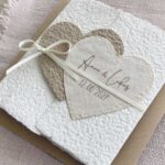Nahaufnahme personalisierte Hochzeitseinladung aus handgeschöpftem Papier im Gatefold Design. Motiv sind zwei ineinanderspielende Herzen in zwei verschiedenen beige-tönen. Titel auf dem Herz ist der Name des Brautpaares und das Hochzeitsdatum. Verschlossen mit einem Satinband in creme.
