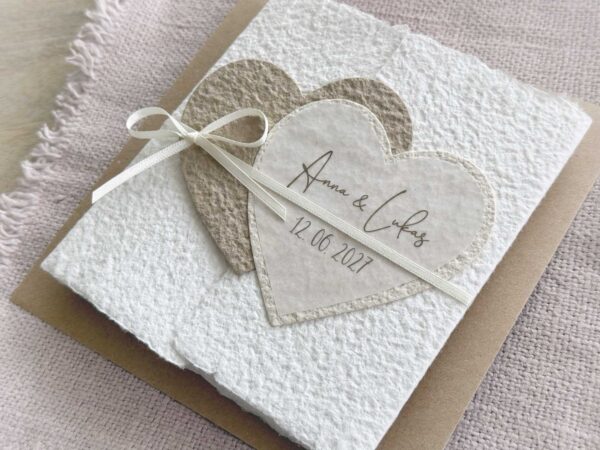 Nahaufnahme personalisierte Hochzeitseinladung aus handgeschöpftem Papier im Gatefold Design. Motiv sind zwei ineinanderspielende Herzen in zwei verschiedenen beige-tönen. Titel auf dem Herz ist der Name des Brautpaares und das Hochzeitsdatum. Verschlossen mit einem Satinband in creme.