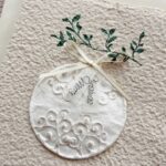 Detailaufnahme Weihnachtskarte Weihnachtskugel aus handgeschöpftem Büttenpapier mit reliefartigem Kugelmotiv, grünem Zweig und cremefarbener Satinschleife.